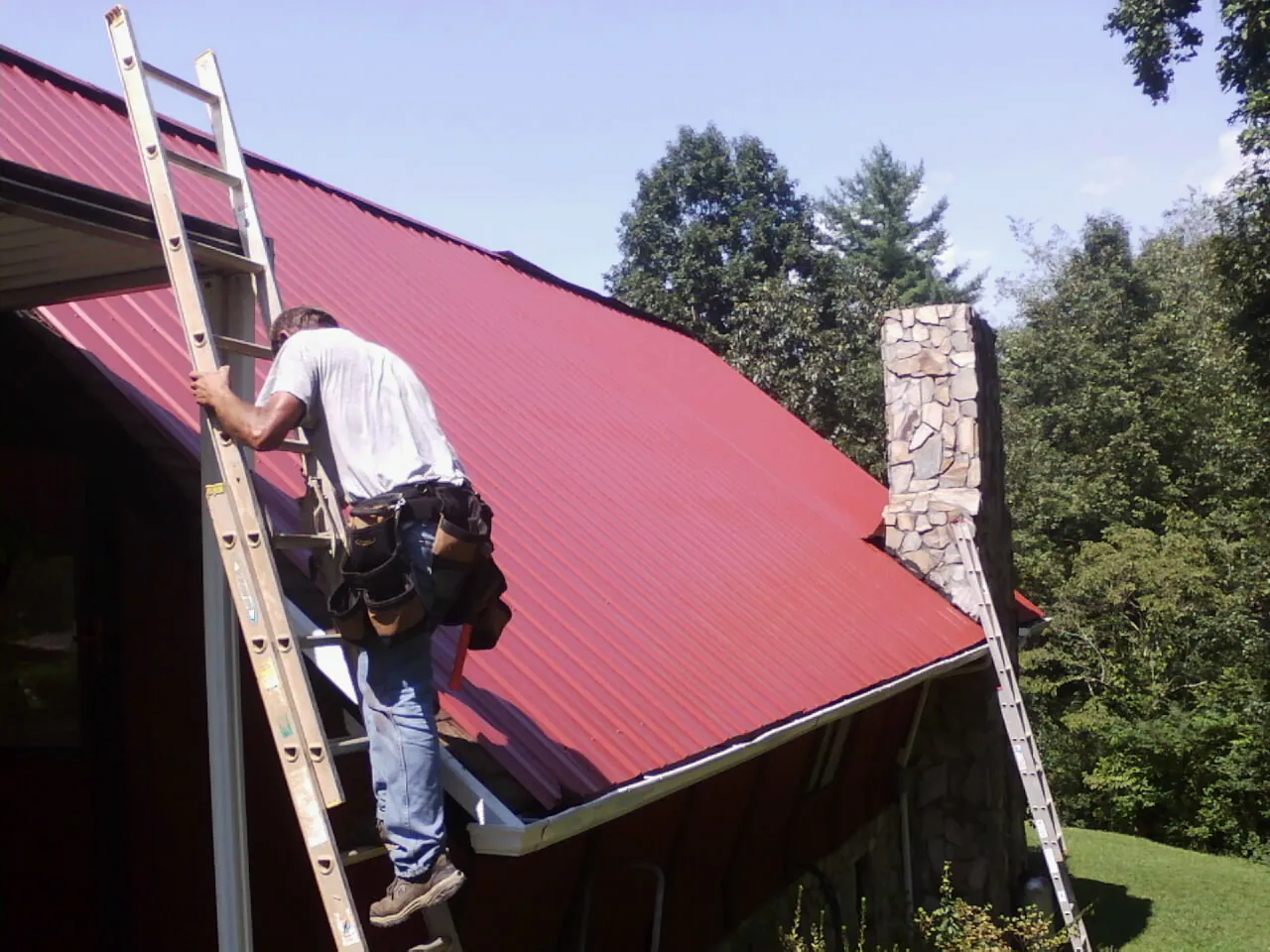 Roofer on a ladder beside a red metal roof and stone chimney — Soffit & Fascia Repair by Roofing Brothers in Stella